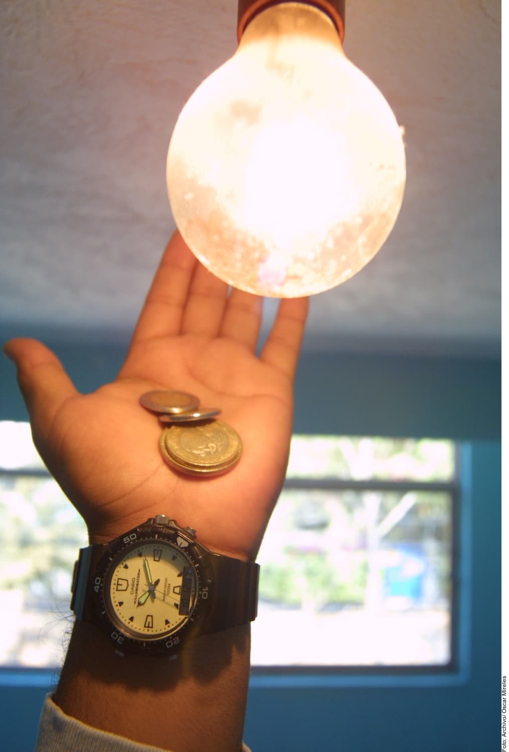 Mano sosteniendo un bombillo encendido con monedas y un reloj de pulsera de fondo.