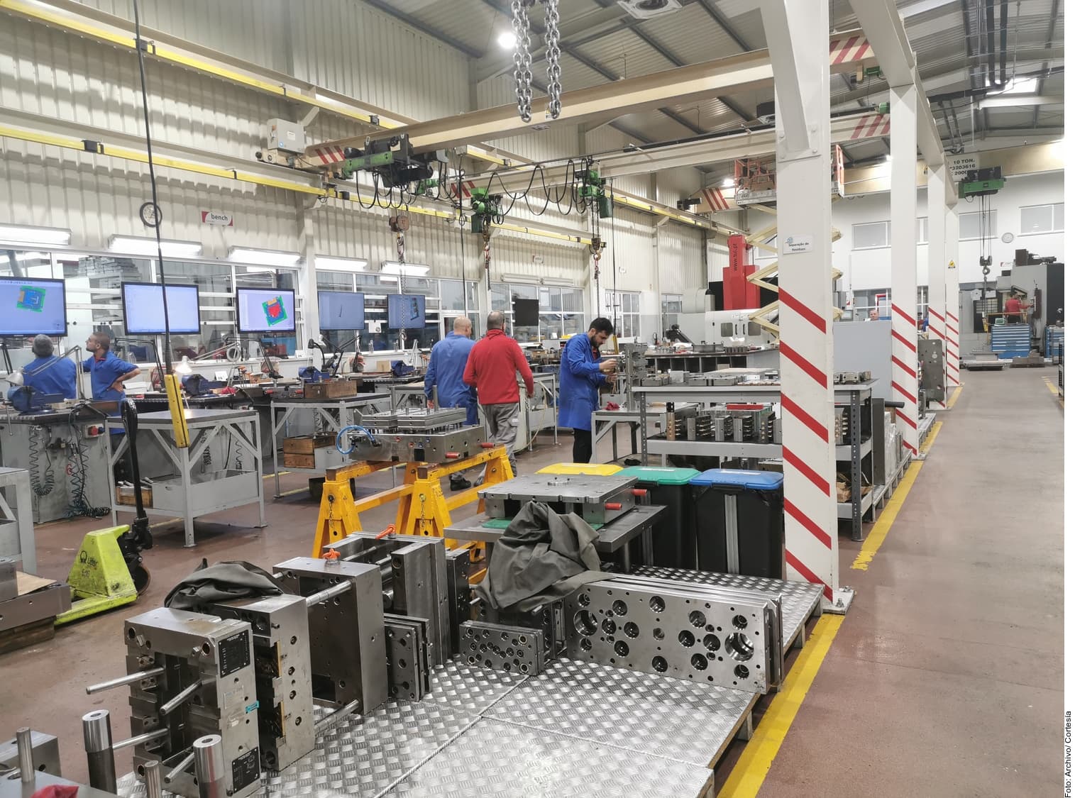 Interior de planta manufacturera con trabajadores en uniforme azul y rojo operando maquinaria industrial y moldes metálicos bajo grúas aéreas.