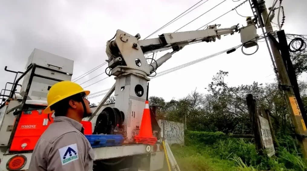 Trabajador de la CFE con casco amarillo operando un vehículo especializado con brazo telescópico cerca de postes y cables de electricidad.