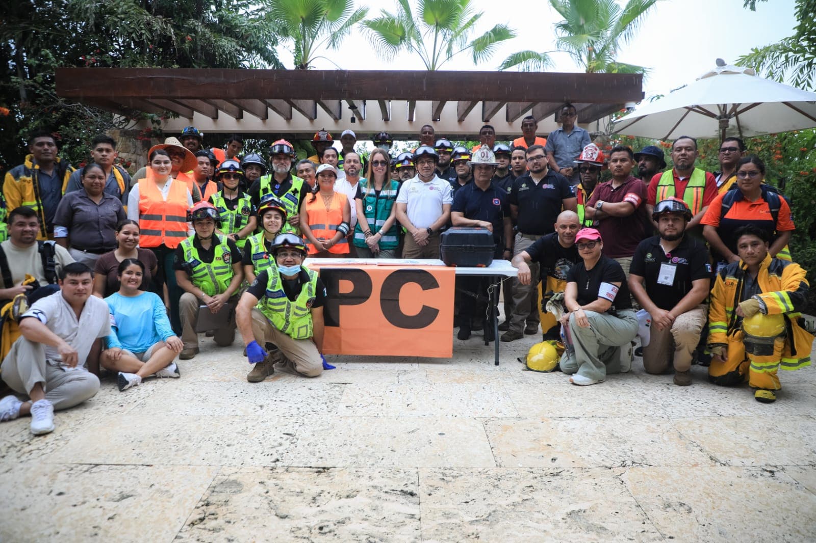 Decenas de personas con chalecos de seguridad naranja y amarillo posando frente a un cartel con las letras 'PC' durante un ejercicio de simulacro bajo una estructura de madera.