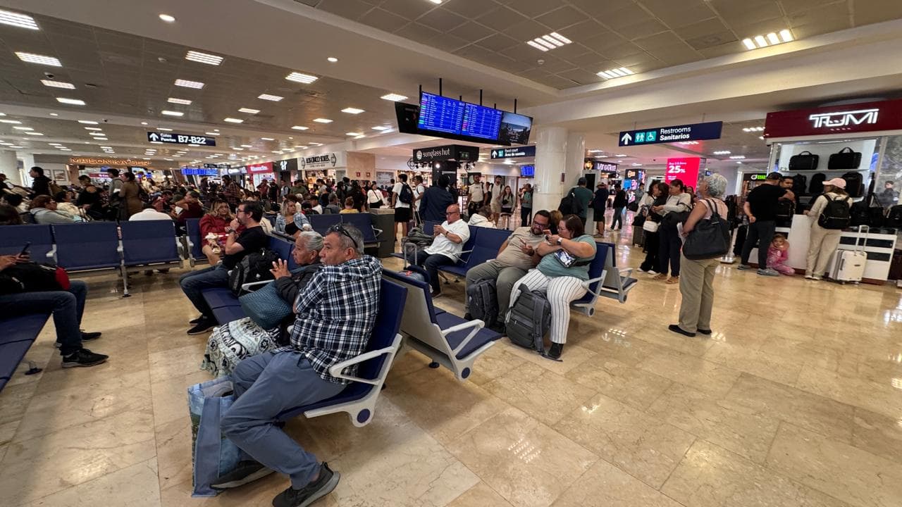 Sala de espera del Aeropuerto Internacional de Cancún con decenas de pasajeros en asientos azules, paneles informativos de vuelos y movimiento intenso de viajeros.