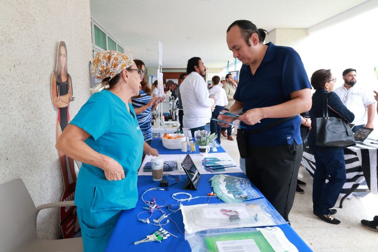 Un hombre en polo azul oscuro atiende a una mujer en turquesa en un stand con materiales médicos durante un evento de salud y bienestar.