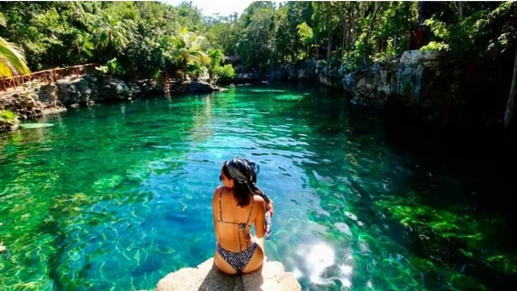 Persona en traje de baño sentada en una roca observando un cenote de agua cristalina y turquesa rodeado de vegetación y formaciones rocosas.