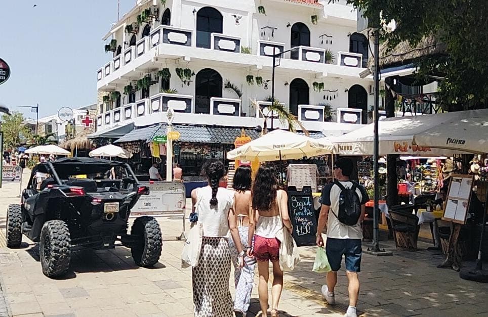 Calle peatonal en Playa del Carmen con comercios, vendedores ambulantes, turistas y un vehículo decorado de fondo, con edificios coloniales blancos al fondo.
