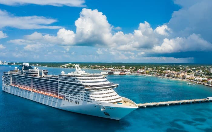 Crucero de gran tamaño atracado en puerto tropical con aguas turquesas y costa con playas y vegetación al fondo.