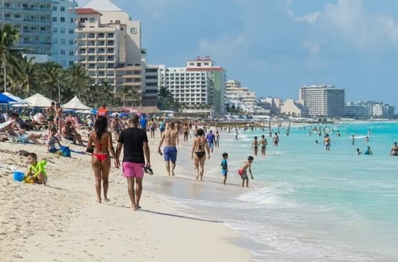 Playa abarrotada de turistas bañándose en aguas turquesas frente a hoteles de gran altura en un destino de playa mexicano.