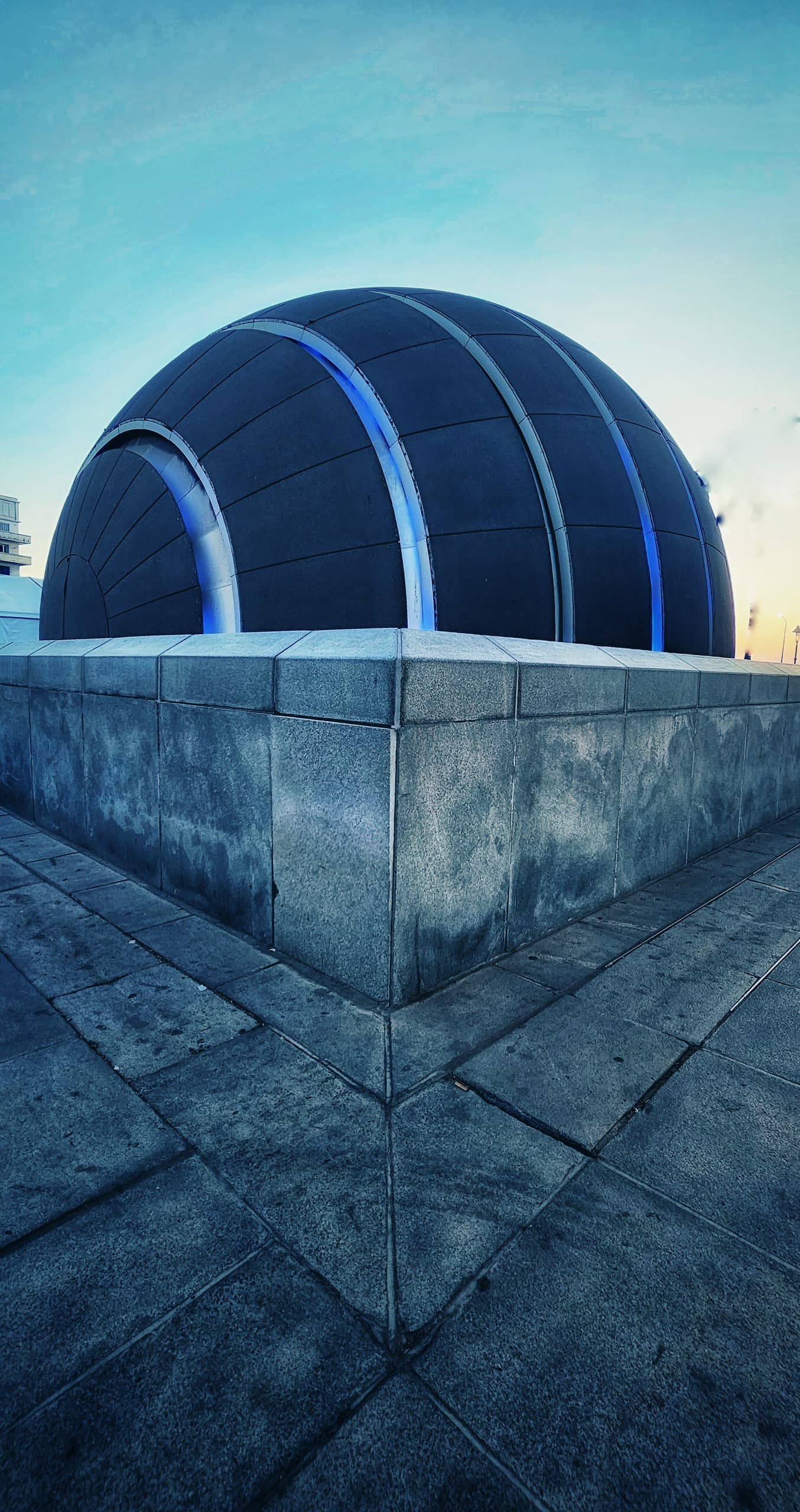 Estructura arquitectónica moderna con forma de cúpula azul oscuro sobre base de concreto, fotografiada contra un cielo despejado.