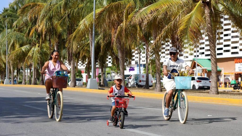 Tres personas en bicicletas circulan por una avenida arbolada de Cancún junto a edificios de apartamentos.