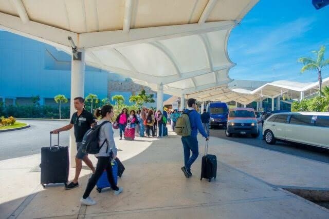Pasajeros con equipaje en la zona de llegadas de un aeropuerto bajo estructura con sombrillas, con vehículos de transporte estacionados al fondo.