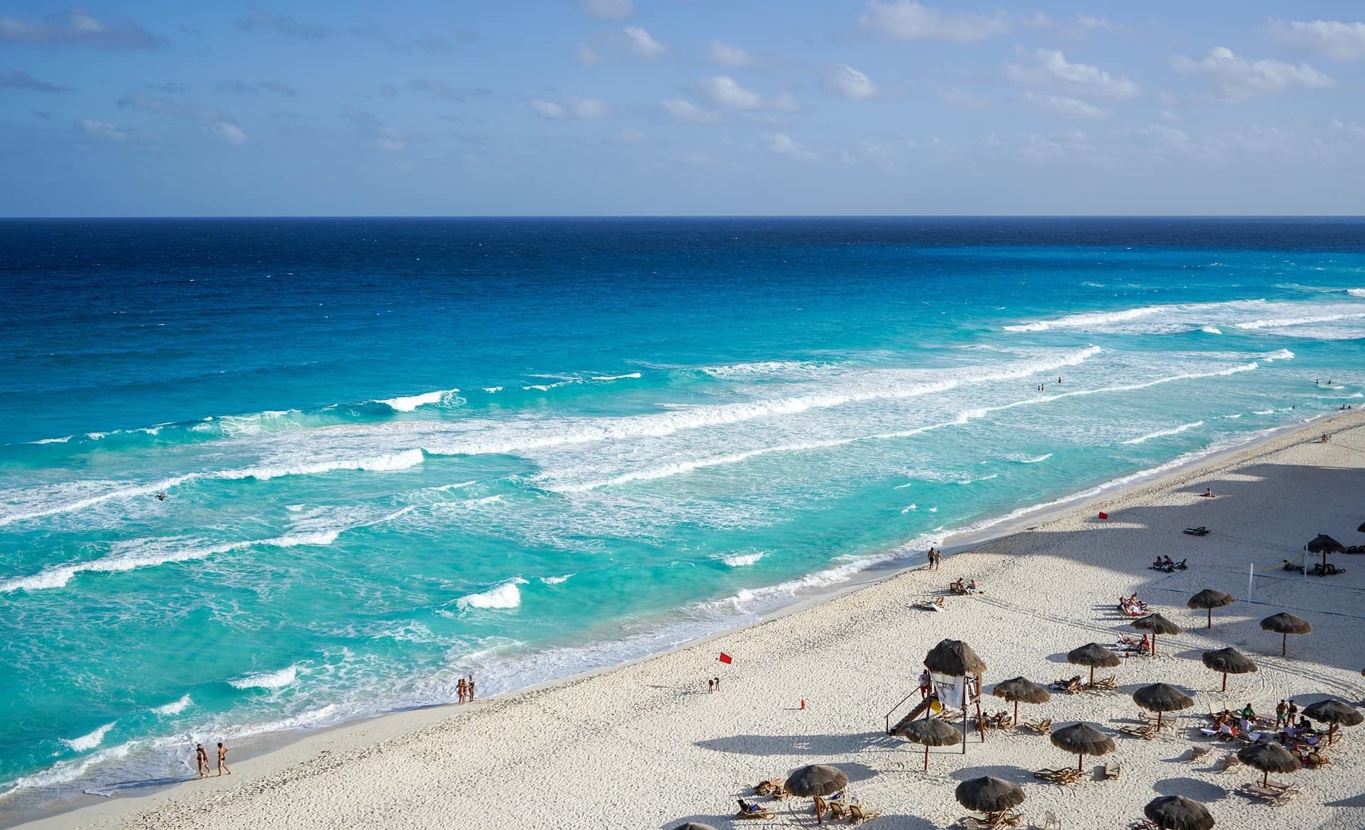 Vista aérea de una playa caribeña con arena blanca, aguas turquesas, olas, sombrillas y bañistas en un día soleado.