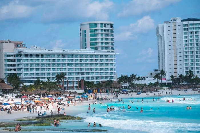Playa con turistas bañándose en aguas turquesas frente a edificios de hotel en zona costera de Quintana Roo.