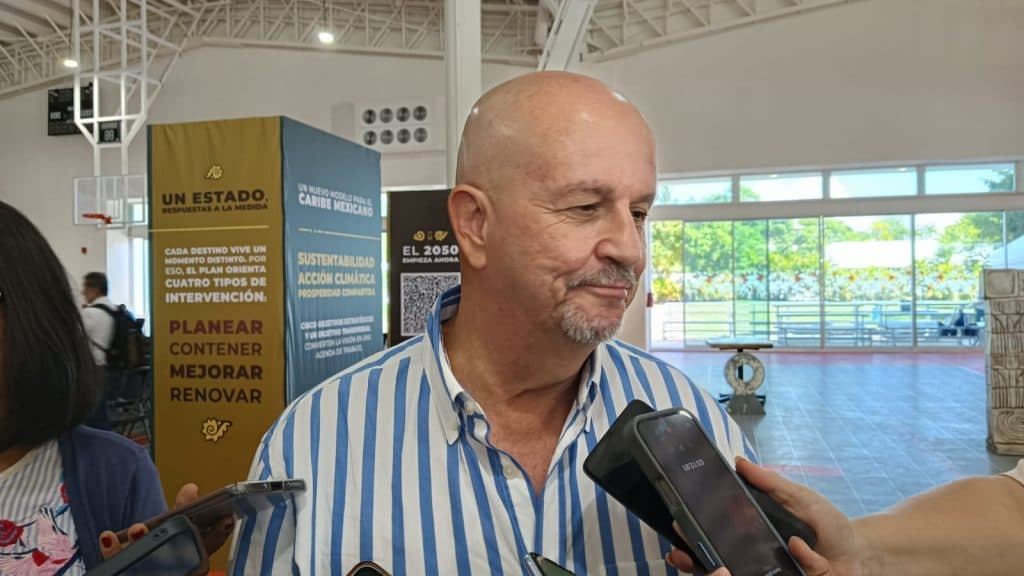 Hombre de mediana edad en camisa de rayas azules y blancas siendo entrevistado por reporteros en un gimnasio.