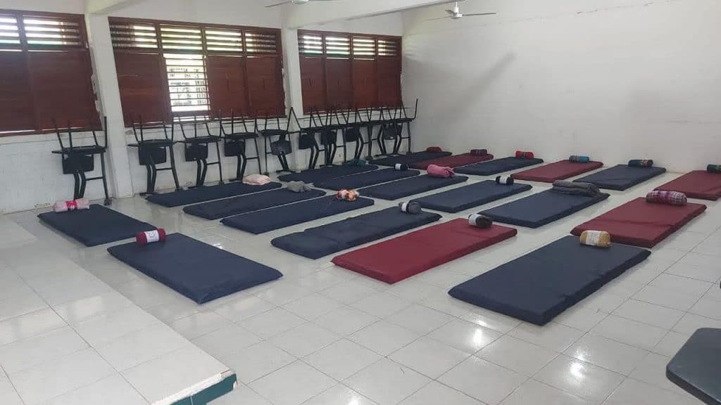 Sala con colchonetas azules y rojas organizadas en filas, junto a sillas apiladas y rollos de tela, en un espacio preparado como refugio temporal.