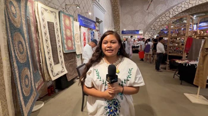 Mujer sostiene micrófono frente a exhibición de textiles y artesanías en una feria o mercado artesanal.