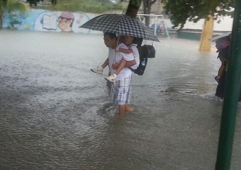 Persona wading through floodwaters while holding an umbrella and a child during heavy rainfall in an urban area.