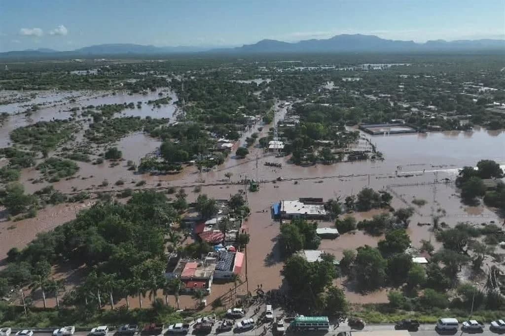 Vista aérea de una zona urbana y rural inundada por agua marrón que cubre calles, casas y campos, con montañas al fondo bajo cielo despejado.