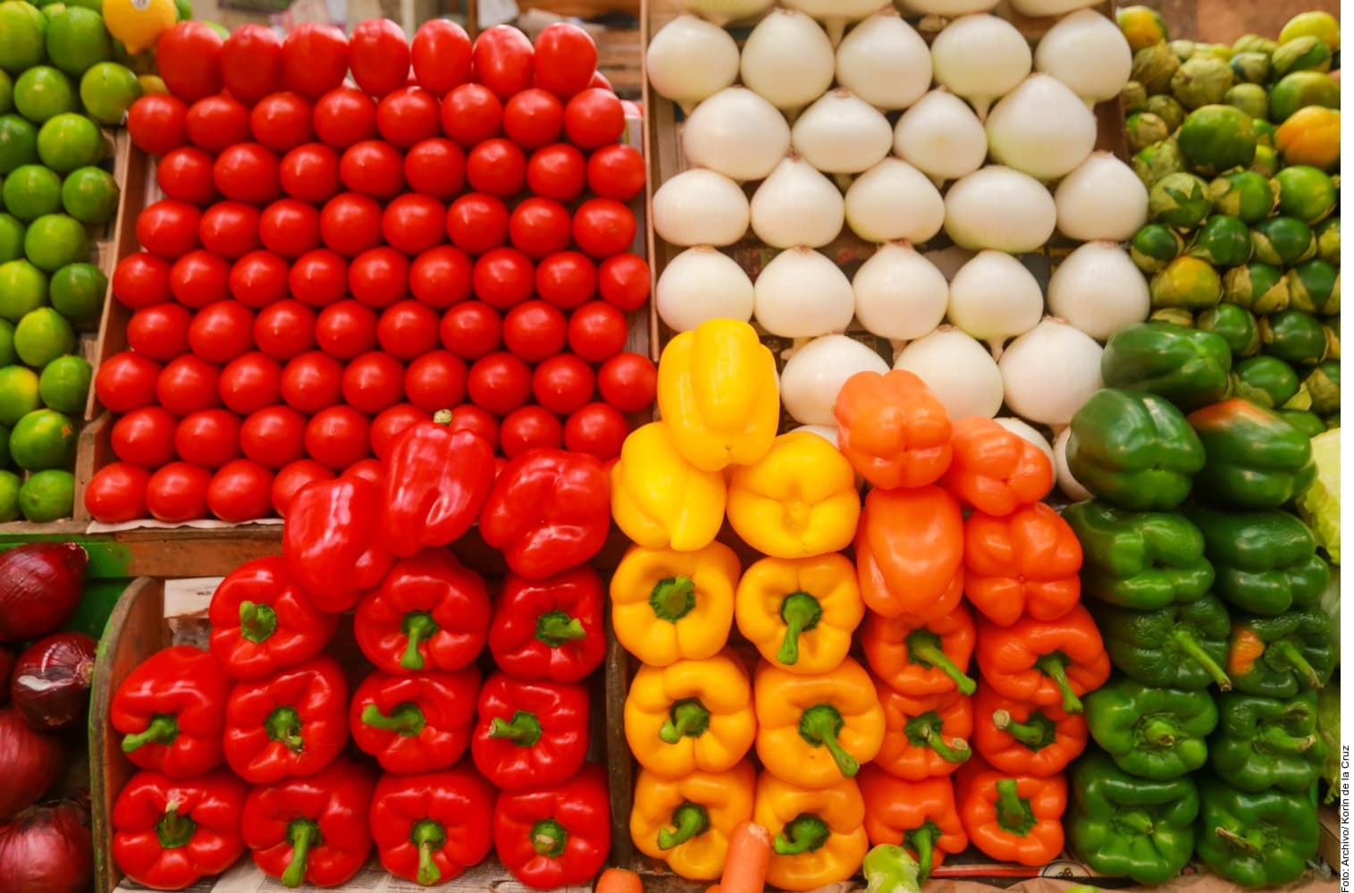 Mercado con puestos de venta de frutas y verduras frescas, incluyendo tomates rojos, pimientos amarillos, naranjas, pepinos verdes, cebollitas y huevos blancos.