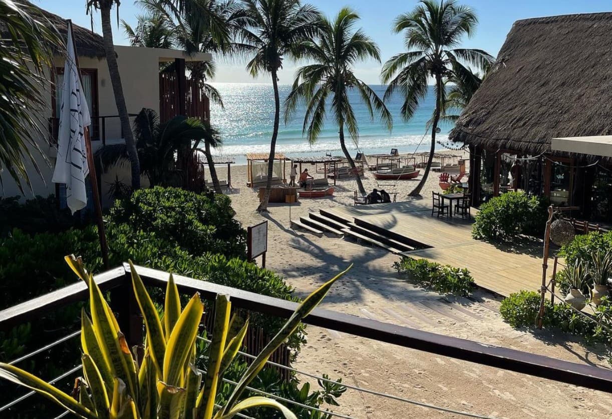 Vista de una playa en Tulum con hoteles sustentables de arquitectura rústica, palapas con techos de palma, palmeras y el mar de fondo.