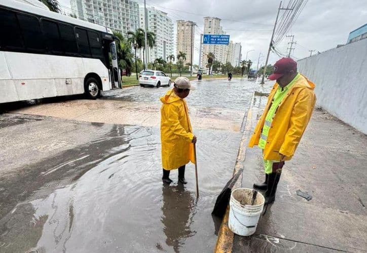 Dos trabajadores con chalecos amarillos y reflectivos drenan agua de una calle inundada en una zona urbana con edificios de fondo durante lluvias intensas.