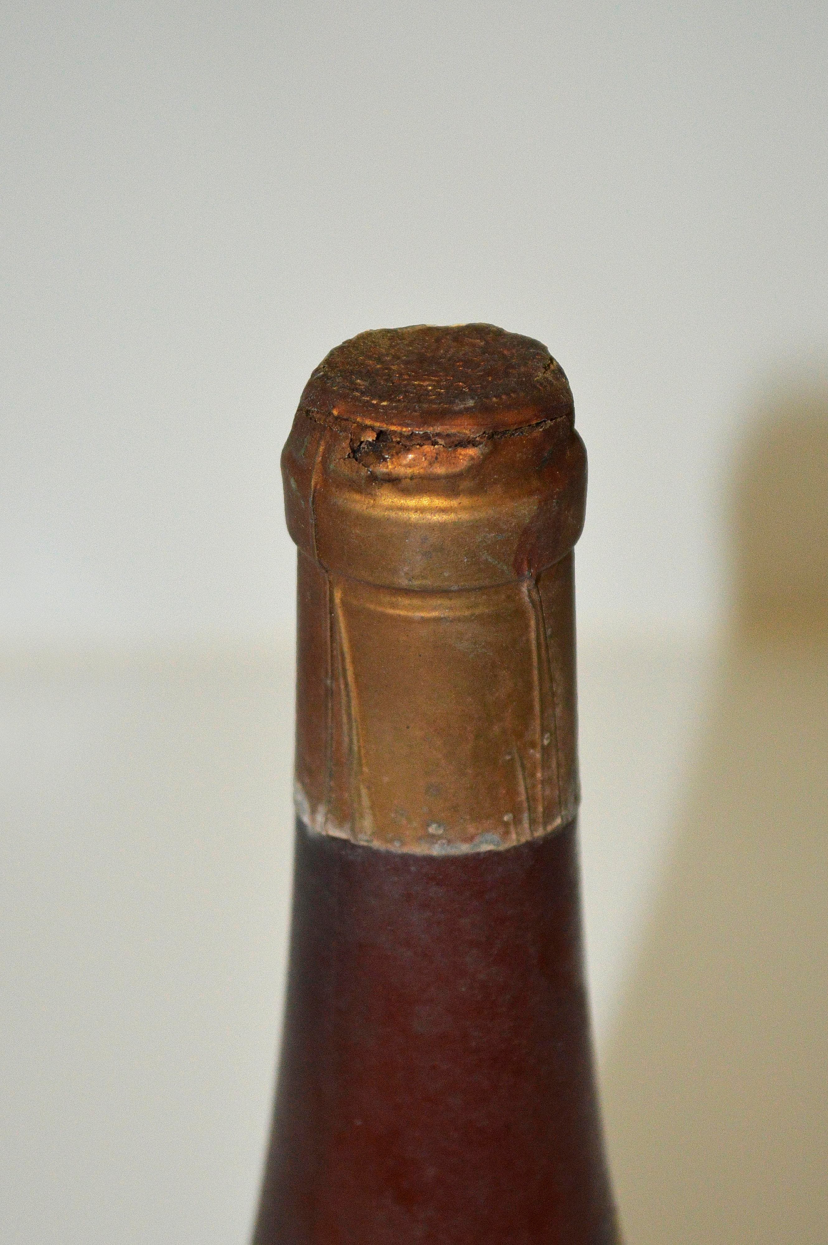 Detalle del cuello y boca de una botella de vidrio oscuro con oxidación en la cápsula metálica superior.