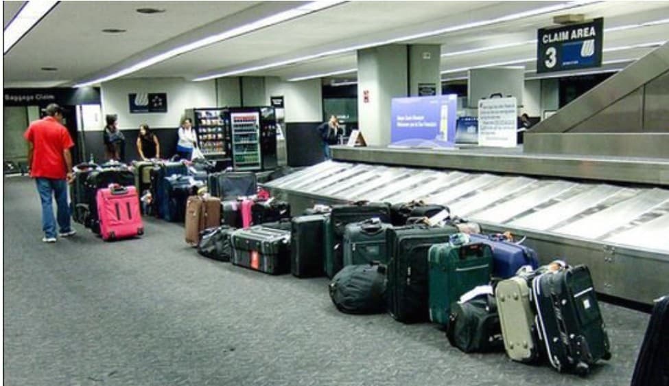 Pasajeros esperan junto a maletas en la cinta transportadora del área de reclamo de equipaje en un aeropuerto.