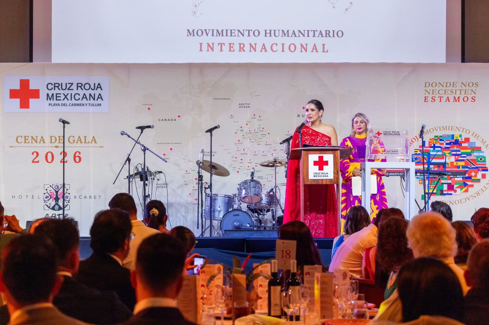 Dos mujeres en el podio durante la Cena de Gala 2026 de Cruz Roja Mexicana en Hotel Xcaret, con escenario, banda en vivo y asistentes al fondo.