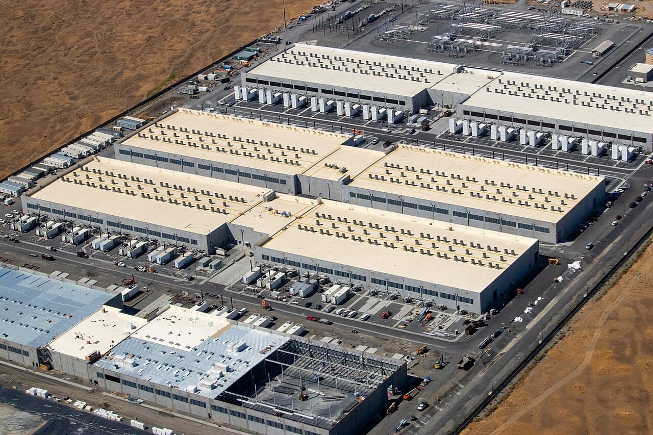 Vista aérea de varios edificios industriales grandes con techos blancos y numerosas unidades de climatización y equipos eléctricos en el techo, junto a una subestación eléctrica y zonas de construcción.
