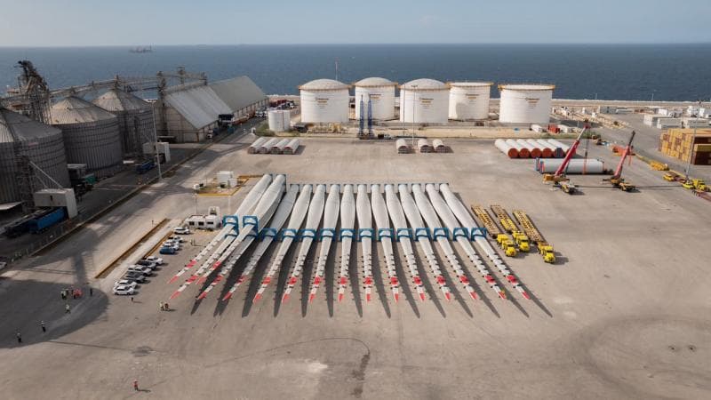 Vista aérea de componentes de turbinas eólicas organizados en el patio de un puerto industrial junto al mar, con silos de almacenamiento y grúas de carga.