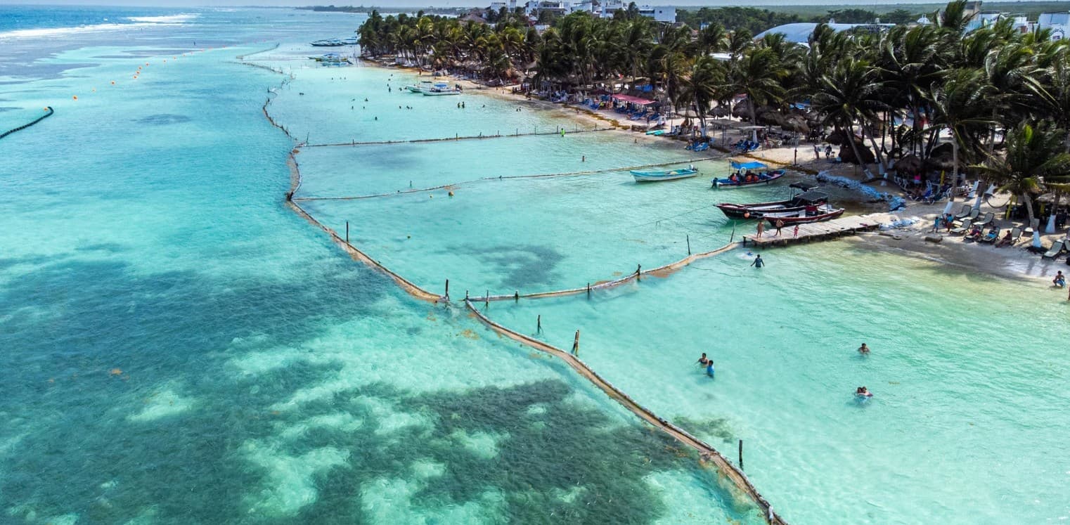 Vista aérea de una playa tropical con aguas turquesas, redes de pesca, embarcaciones tradicionales y bañistas en la arena, con edificios y palmas de coco al fondo.