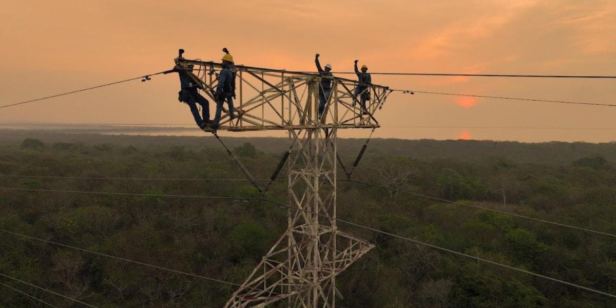 Trabajadores realizan mantenimiento en una torre de transmisión eléctrica de alta tensión al atardecer, con bosque de fondo.