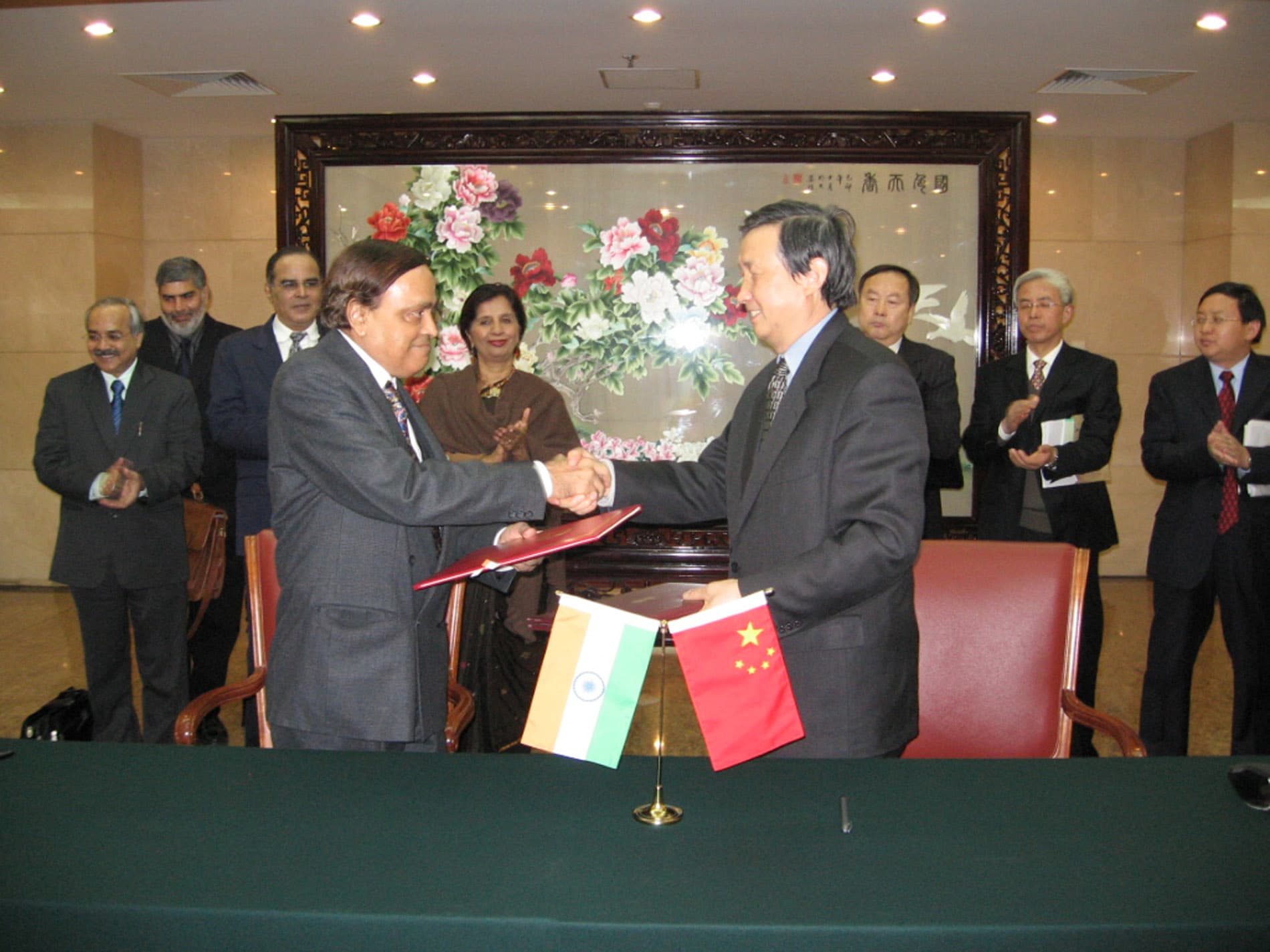 Dos funcionarios de petroleras estatales estrechan manos durante la firma de un acuerdo de cooperación, con banderas de India y China visibles.