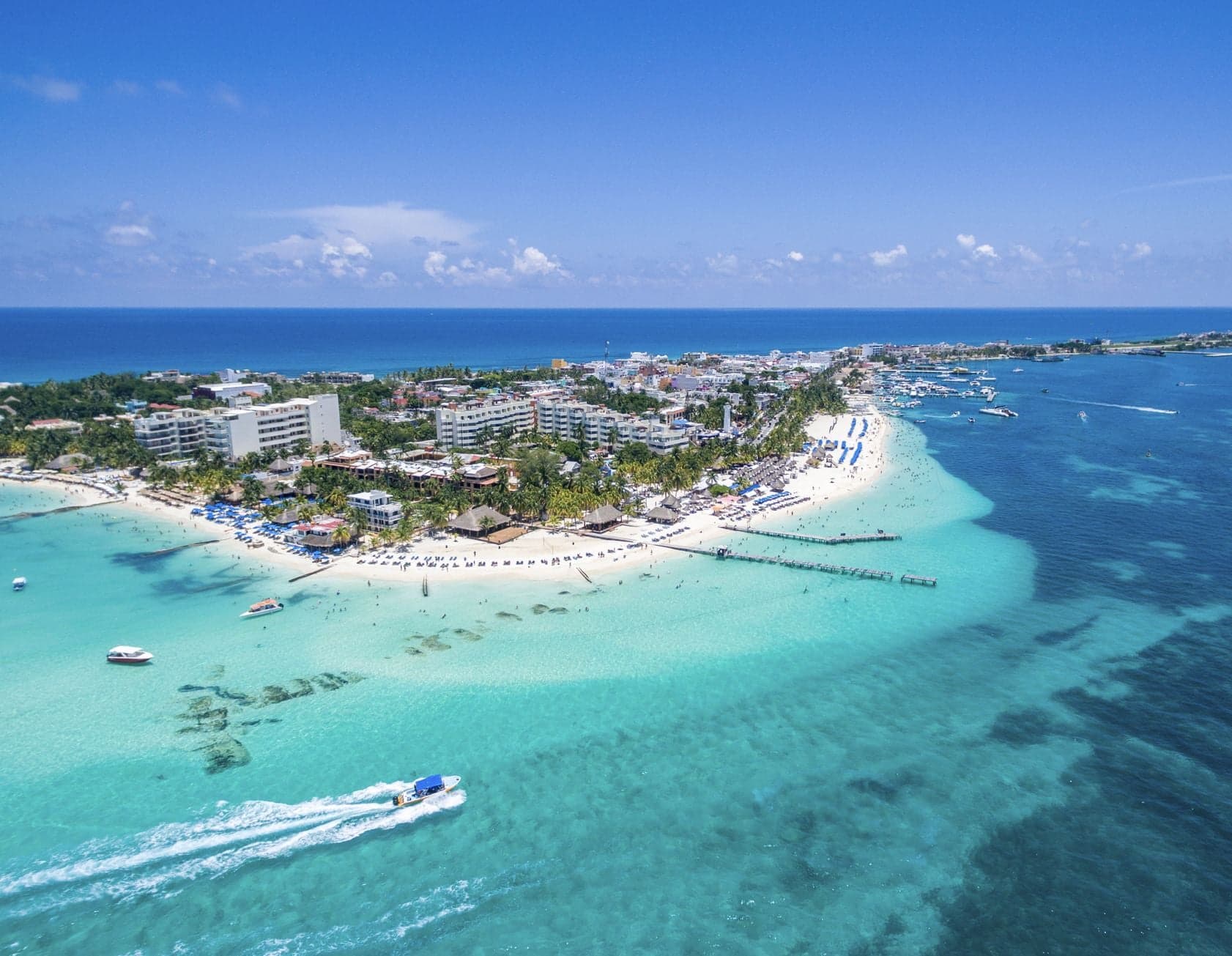 Vista aérea de una playa tropical con aguas turquesas cristalinas, arena blanca, embarcaciones y desarrollo hotelero en la Riviera Maya.