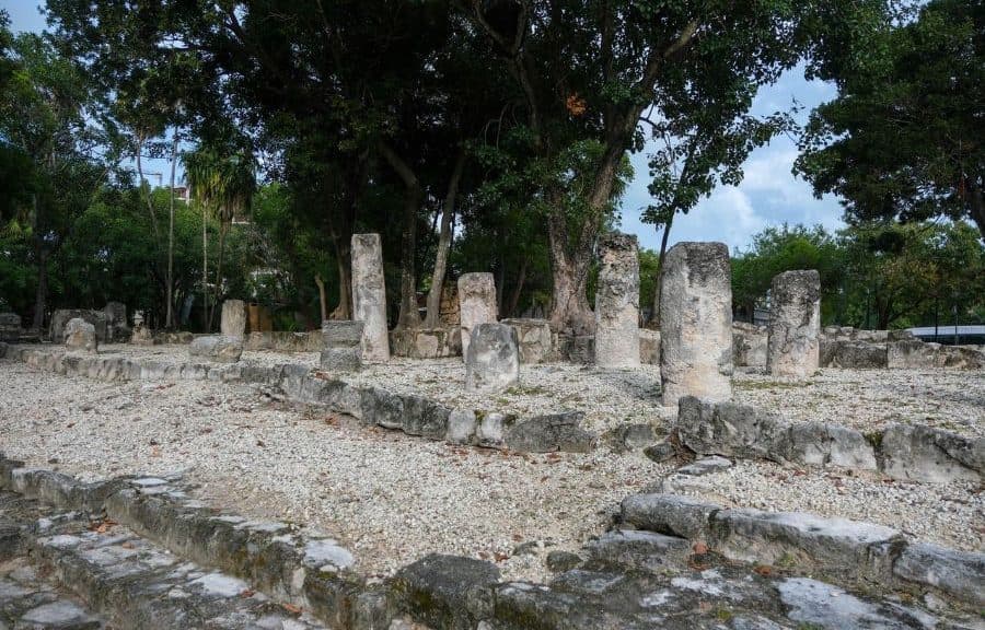 Ruinas arqueológicas mayas con columnas de piedra y vegetación tropical de fondo bajo cielo despejado.