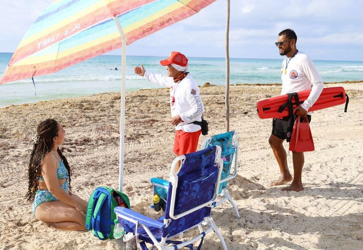 Guardavidas en uniforme blanco y rojo interactúan con una bañista en la playa bajo sombrilla de colores, con el mar de fondo.