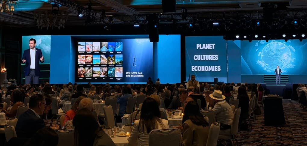 Conferencia de turismo en salón de hotel con cientos de asistentes, presentador en escenario y pantallas mostrando contenido sobre planeta, culturas y economías.