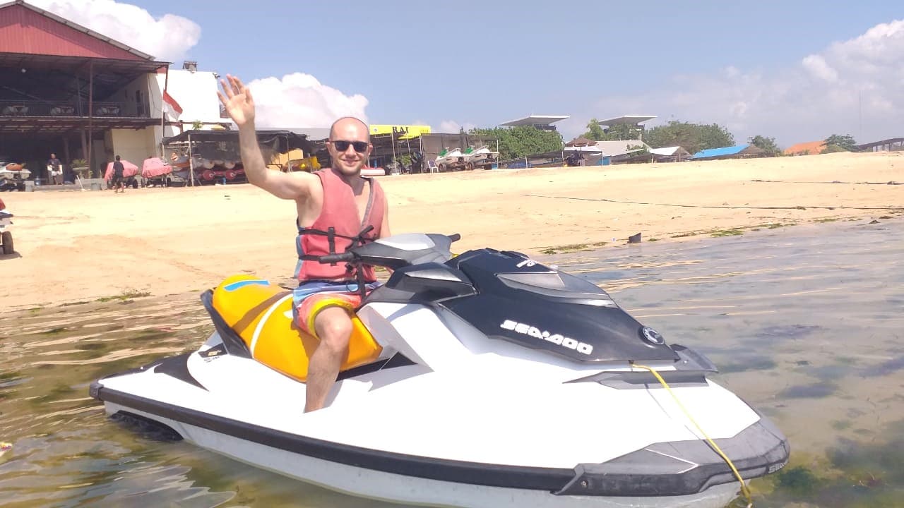 Hombre con chaleco salvavidas waving desde una moto acuática amarilla y blanca en aguas costeras, con instalaciones turísticas de fondo.