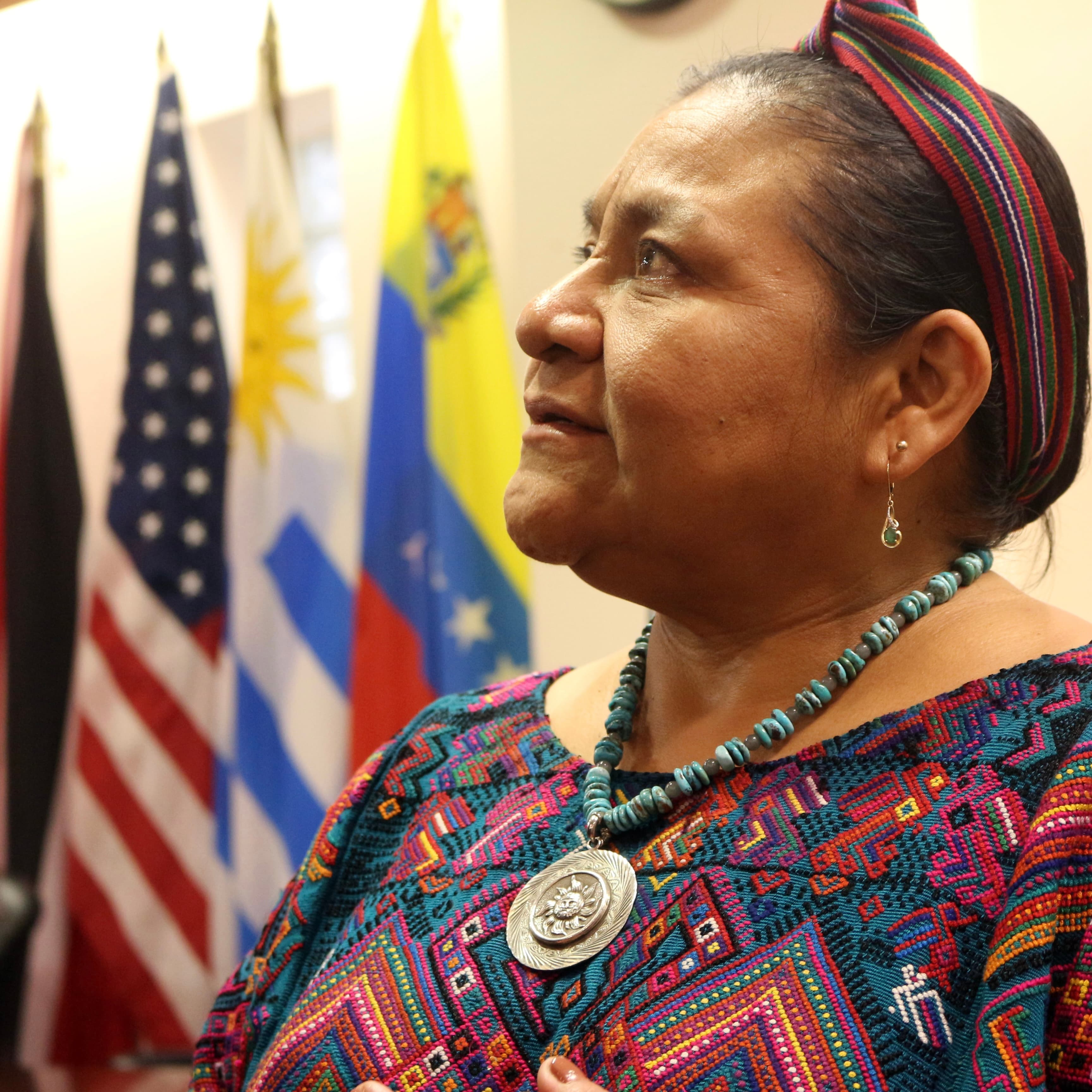 Mujer indígena con huipil bordado de colores, faja tejida en la cabeza, collar de cuentas turquesa y medallón de plata, mirando hacia arriba frente a banderas de varios países.
